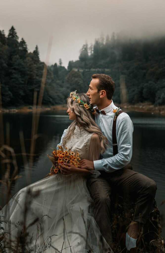 Young groom gently embracing bride with flower wreath and bouquet while sitting on river shore against woods and looking away