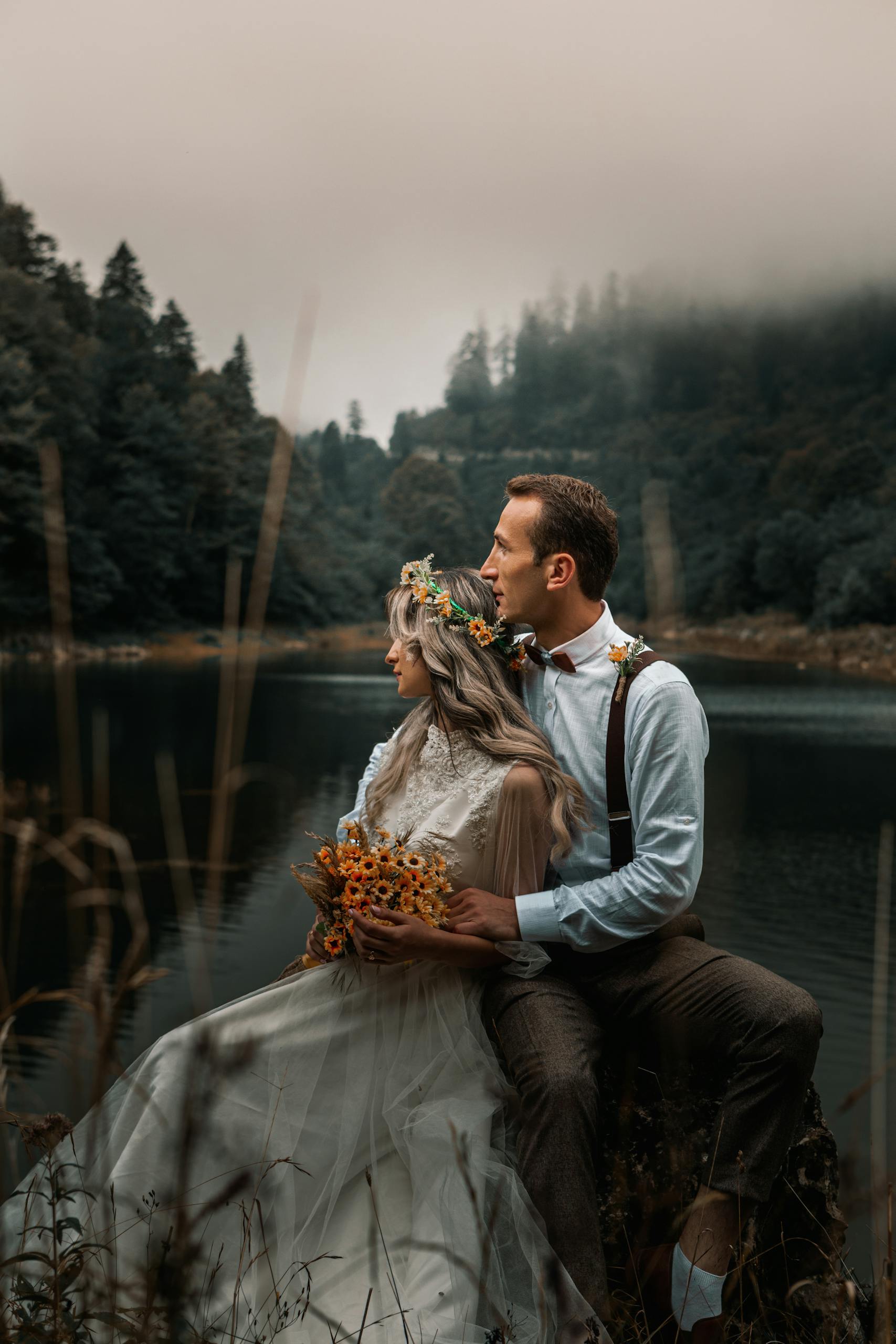 Young groom gently embracing bride with flower wreath and bouquet while sitting on river shore against woods and looking away